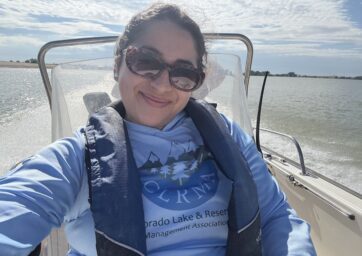 Dominique Garcia sits on a small boat wearing sunglasses and a life vest, smiling while conducting water sampling on a sunny day at a Colorado reservoir.