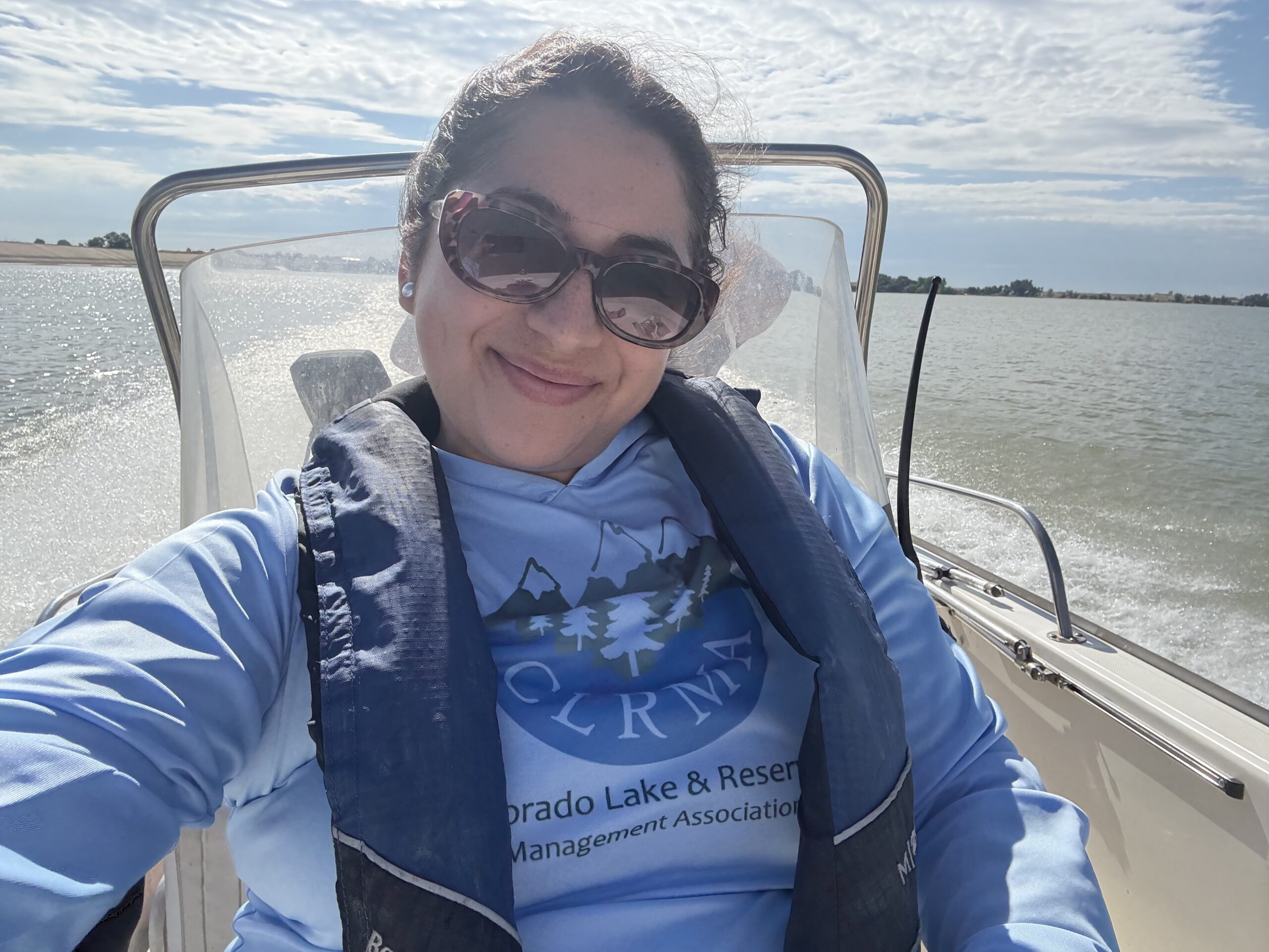 Dominique Garcia sits on a small boat wearing sunglasses and a life vest, smiling while conducting water sampling on a sunny day at a Colorado reservoir.