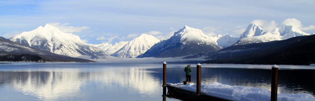 Photo. An individual standing on the end of a pier jutting into a body of water, with snowcapped mountains in the distance.