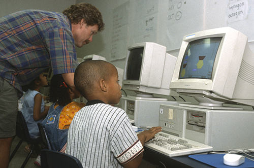 Students working on old computers while a teacher assists