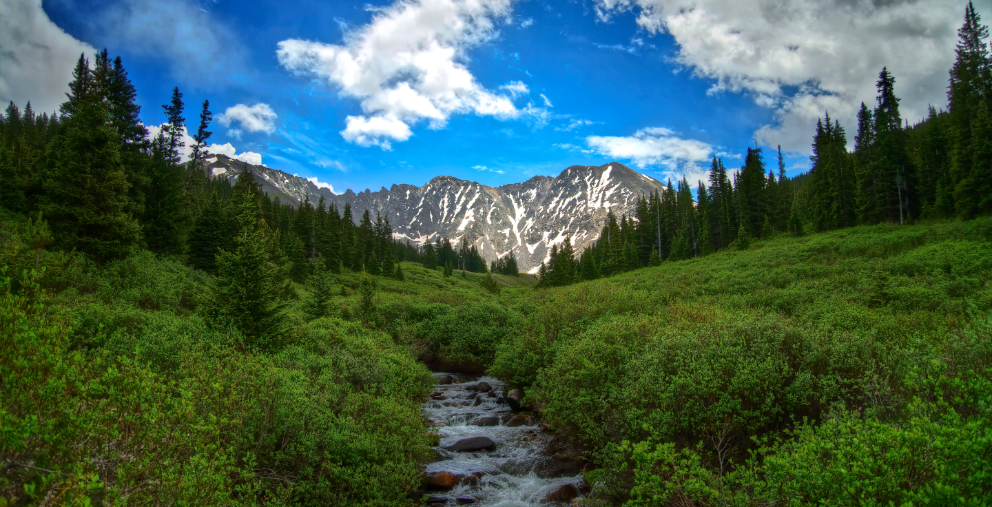 Photo. A creek cuts through green grass and trees with mountains in the distance, a partly cloudy sky overhead.