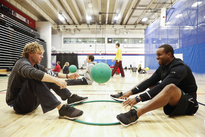 Photo of two adult students sitting on the floor in a gym with a hula hoop on the floor between them and children in the background.