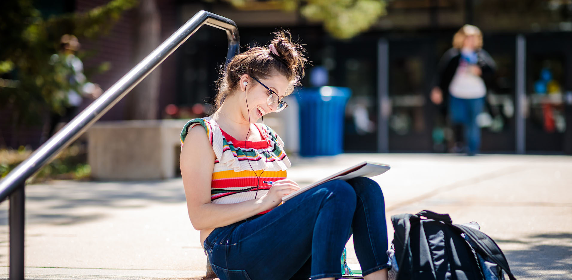 Student sitting on outside stairs writing in notebook and wearing headphones