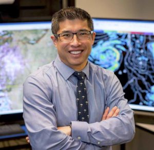 Dr. Sam Ng standing in front of two computer monitors displaying weather maps