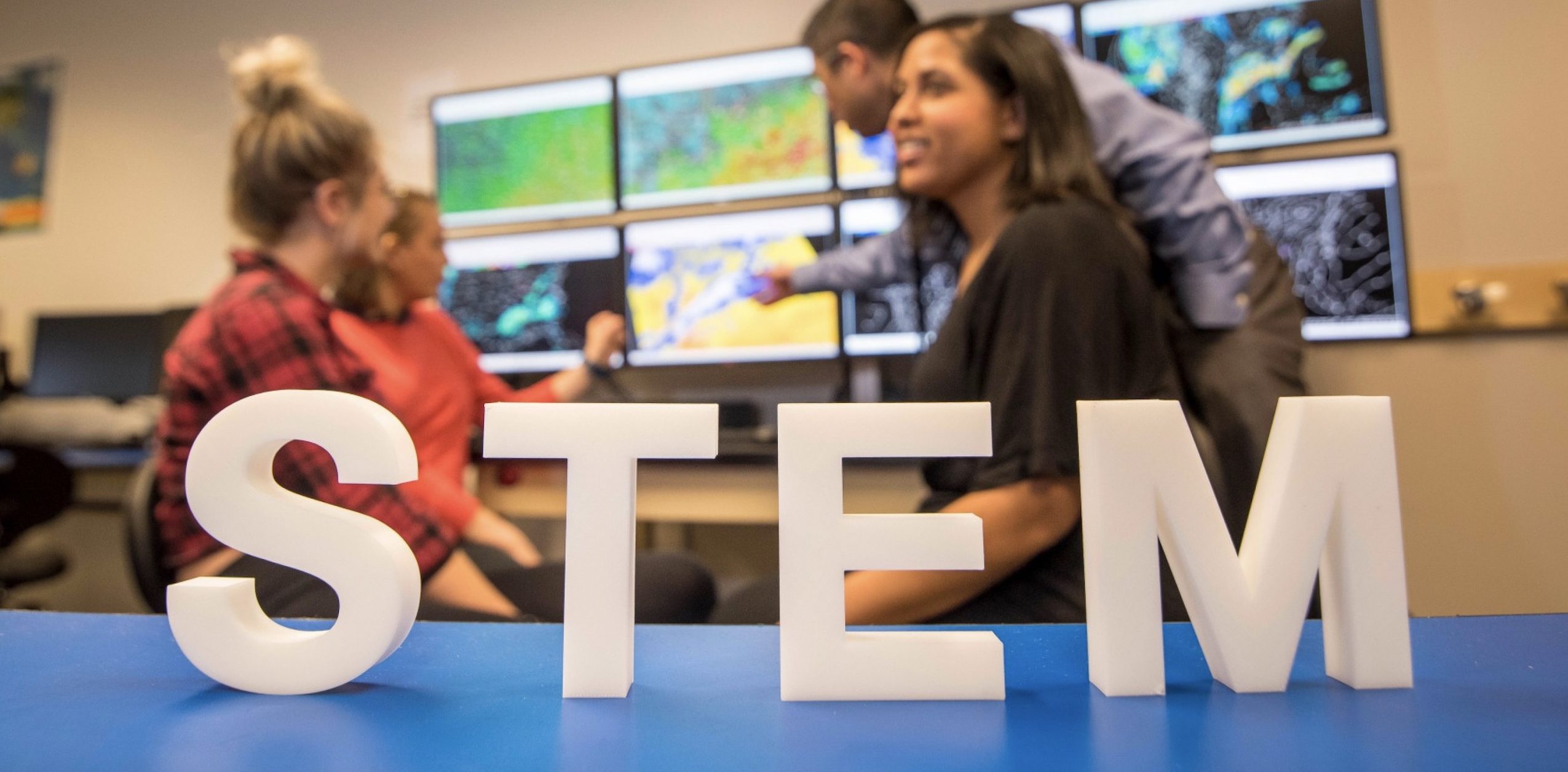 STEM sign on a blue table with three female students and a male instructor behind it discussing weather on computer displays