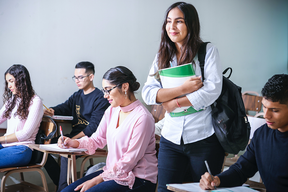 Students in class, one standing with a backpack and notebook