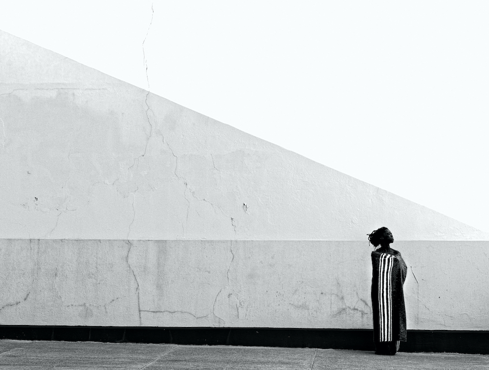 Black and white photo of a person in striped clothing standing against a large, cracked wall with a triangular section.