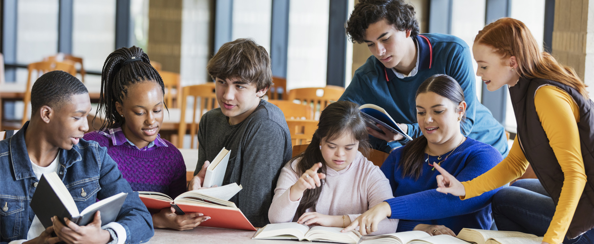 Image of a diverse group of middle school and high school studets sitting together at a table in a library, studying from textbooks and interracting with one another..