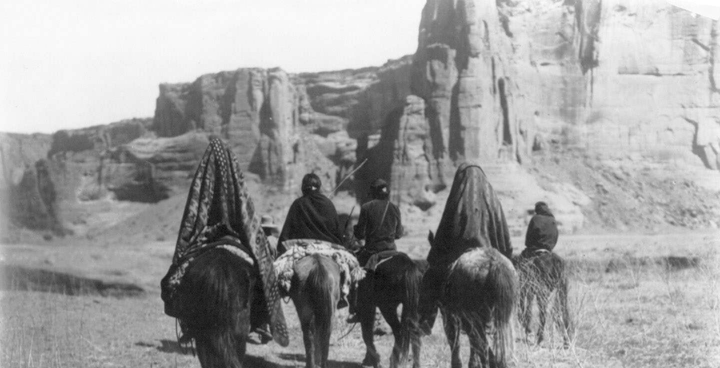 Rear view of Navajo Indians on horseback making their way over the sparse, dry, grassy floor of Tesacod Canyon.