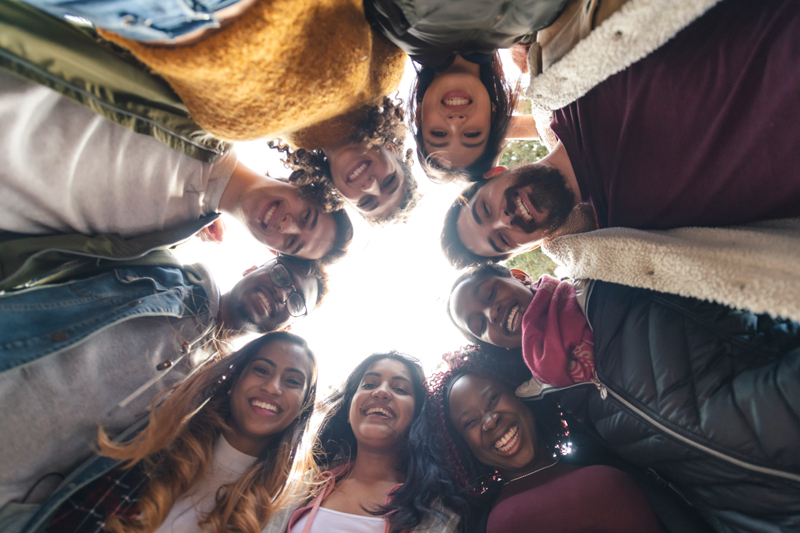 Image of a diverse group of college aged students standing in a circle looking down and smiling at a camera in between them all.