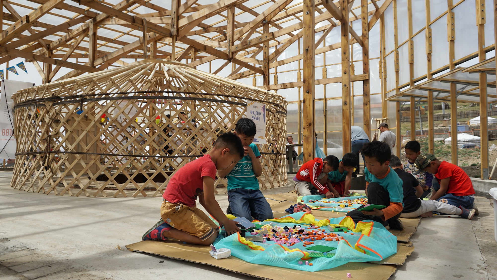 A group of children sit on the floor of a wood-framed and glass-clad structure. The children are interacting with colorful materials.