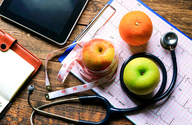 Apples and oranges wrapped around stethoscope and measuring tape on wooden desk.