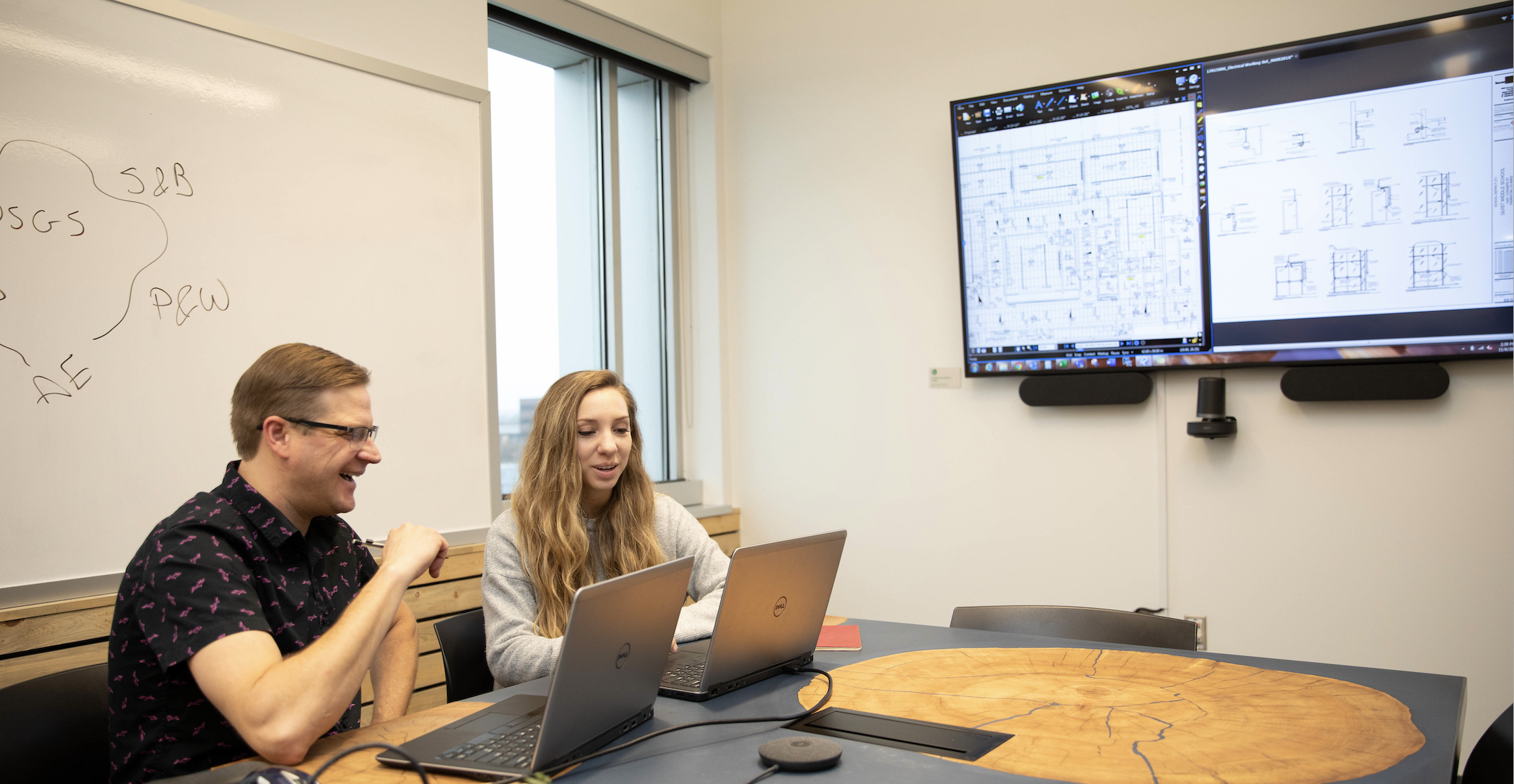 Two people sitting in a conference room at laptops with screens behind