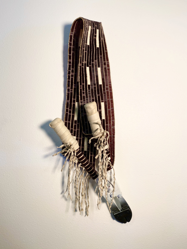 Beaded maroon belt with beige patterns and fringe, integrated with two handles wrapped in beads and feathers hanging on a light wall.
