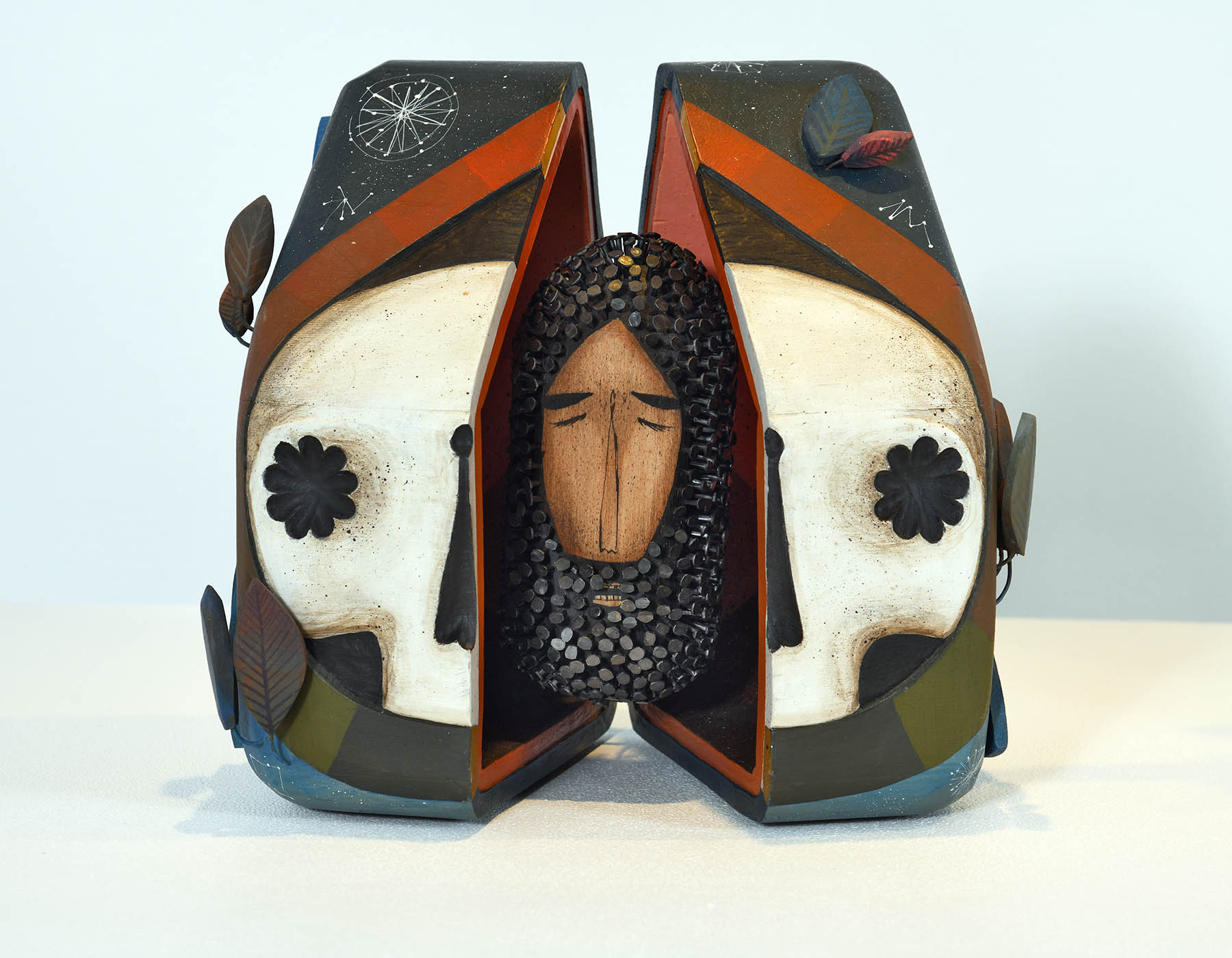 A sculpture of two stylized skull faces flanking a central wooden-textured figure with closed eyes and dark curls.