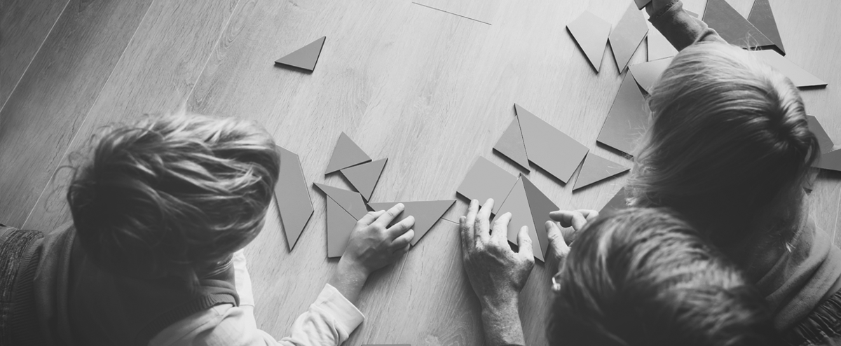 overhaed view of an adult and two children playing with tangrams on a wood floor.