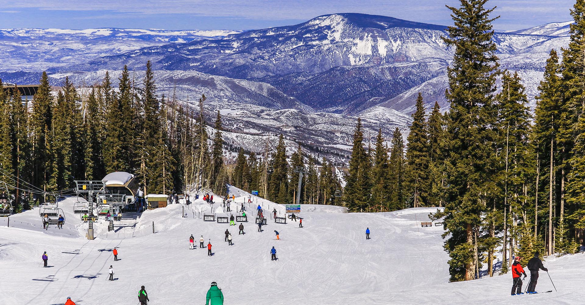Looking down a ski slope from the top with a bunch of skiers and well-worn trails. A scenic vista is the backdrop, truly purple mountain majesty yawning out panoramically, evoking a sense of wonder and awe.