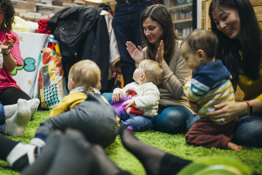 Mothers and their baby children are in a sensory play group. They are singing and dancing together in a circle on the floor.