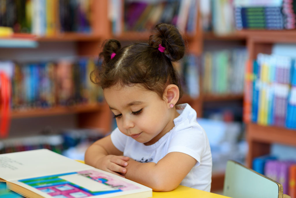 Young girl sitting at a table in a classroom reading a book.