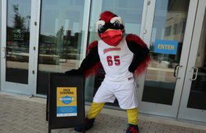 MSU Denver mascot, Rowdy, wearing a mask and posing next to mask mandate sign.