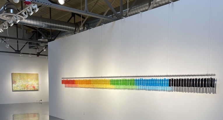A contemporary art gallery features a wall installation of ice pops arranged in a color gradient, with a framed artwork hanging on an adjacent wall.