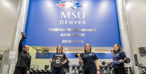 People posed with workout equipment in the gym below the MSU Denver's Human Performance and Sport Athletics sign.