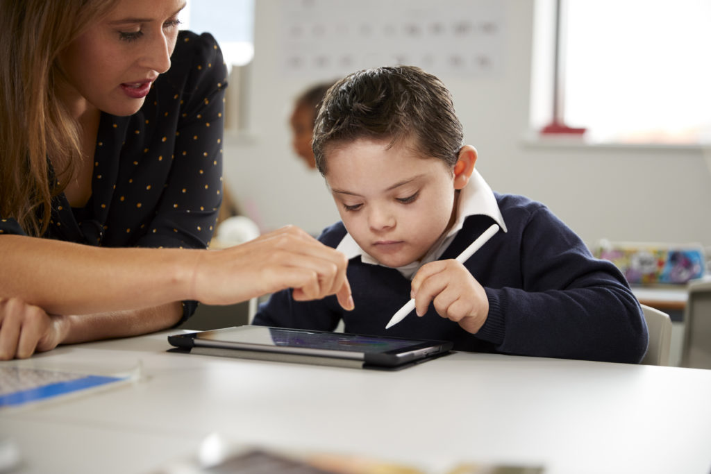 Young female teacher working with a Down syndrome schoolboy sitting at desk using a tablet computer in a primary school classroom, front view, close up