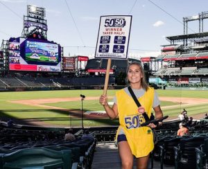 A girl selling 50/50 Raffle tickets stands behind home plate at Coors Field with the field behind her
