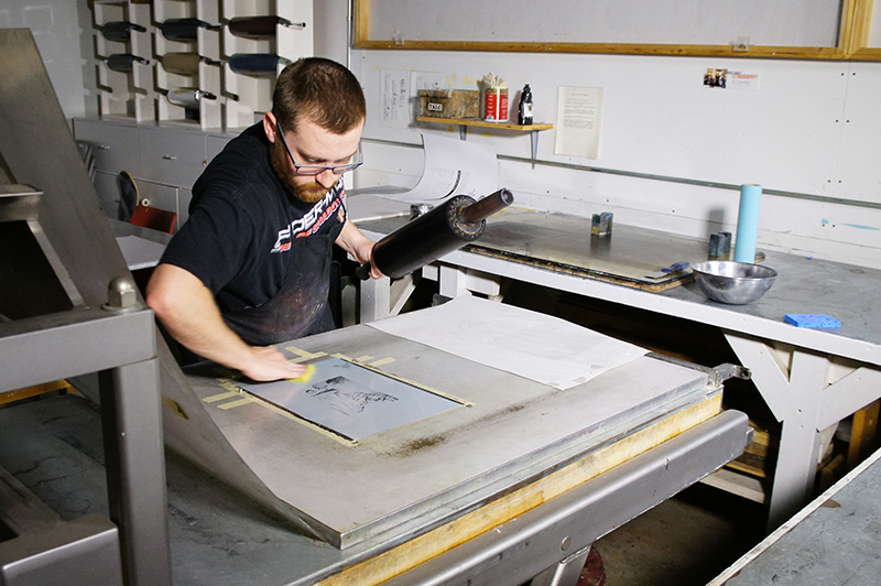 A man using a brayer to ink a metal printing plate in a printmaking studio.