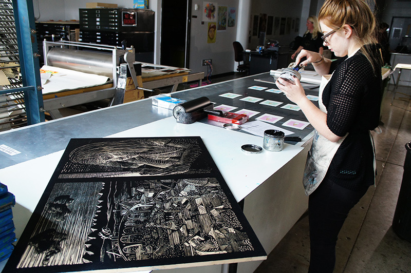 A person works on printmaking projects in an art studio, surrounded by tools and a carved woodblock with landscape and contemplative figure on a drafting table