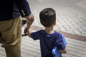 A parent holding a child's hand; both photographed from behind.