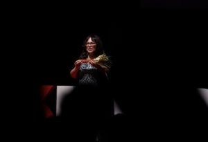 A woman with glasses holds a piece of dried corn in both hands with a dark background.