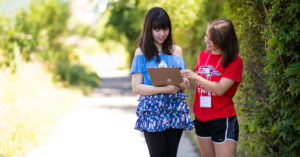 Two women interact with one holding a clipboard and the other pointing on the clipboard.