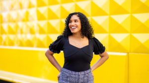 Ines Calvete Barrio in front of a bright yellow wall, her hands on her hips and something happening off to the left that must be making her very happy, evidenced by a glowing smile.