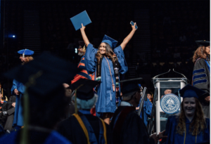 An excited graduate in cap and gown standing arms outstretched with a just-recieved diploma cover in hand.