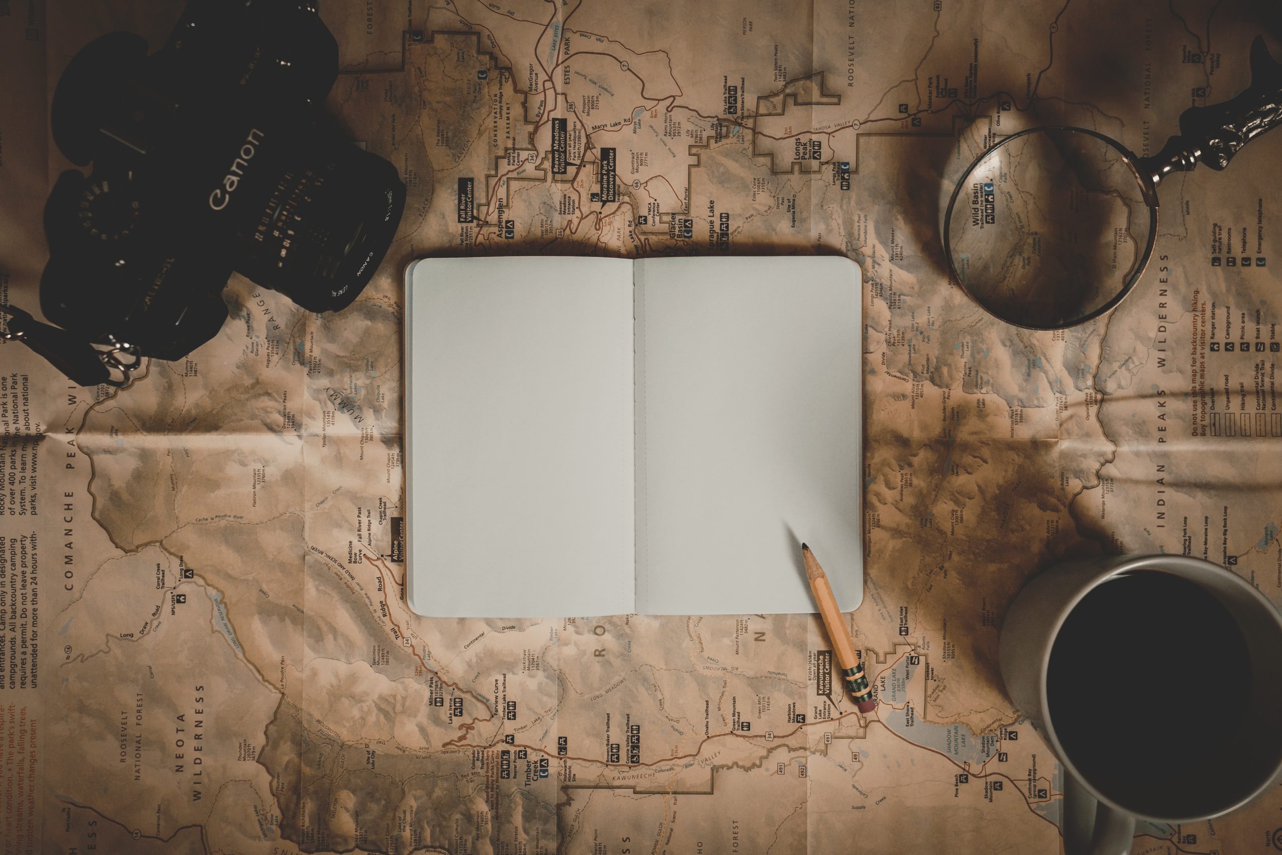 A canon camera, a blank open notebook, a pencil, coffee mug and magnifying glass all sitting on a spread out map
