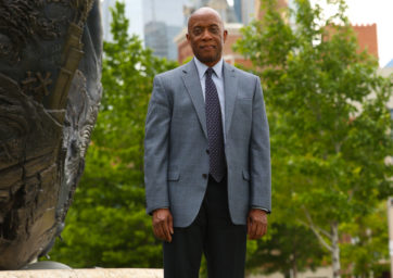 Russell Noles standing next to the statue in front of the Student Success Building with the Tivoli and trees in the background.