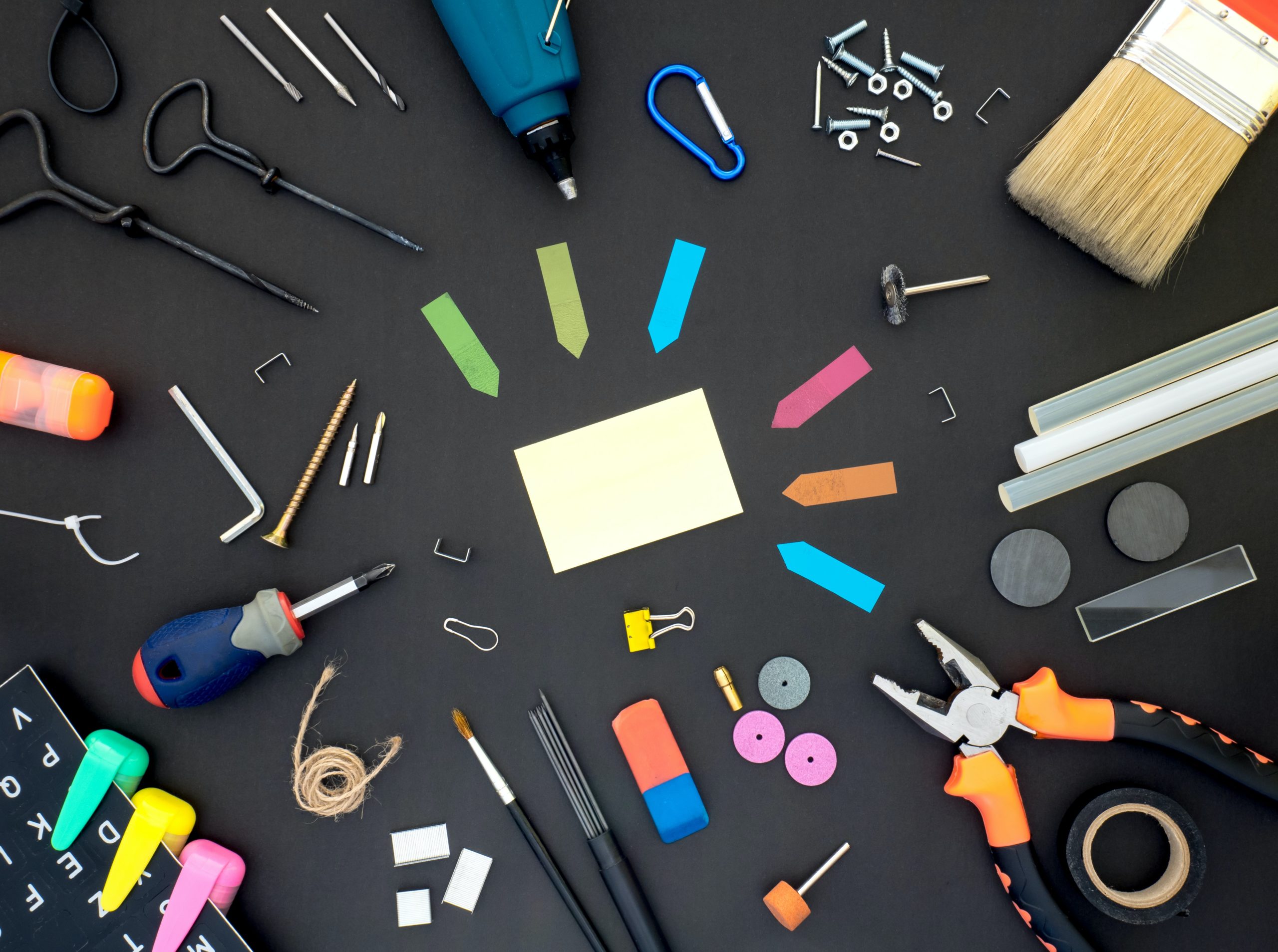 An assortment of tools laying on a black surface including post-its, screwdrivers, wrenches, tape, paintbrushes, highlighters, glue sticks, staples, magnets, and paperclips.
