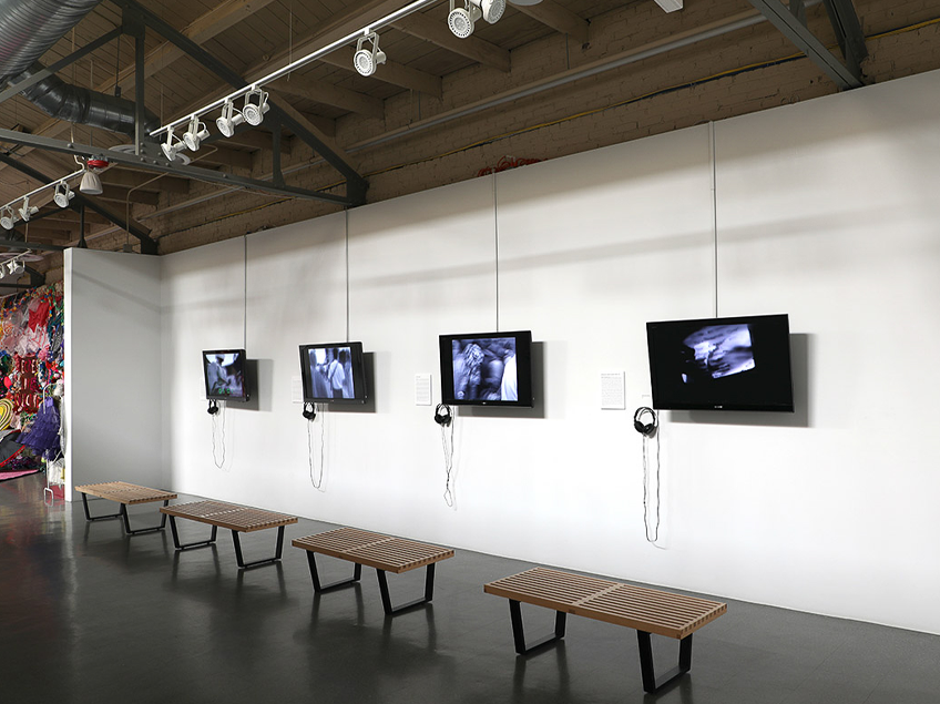 Art gallery with five mounted monitors, wooden benches, and headphones.
