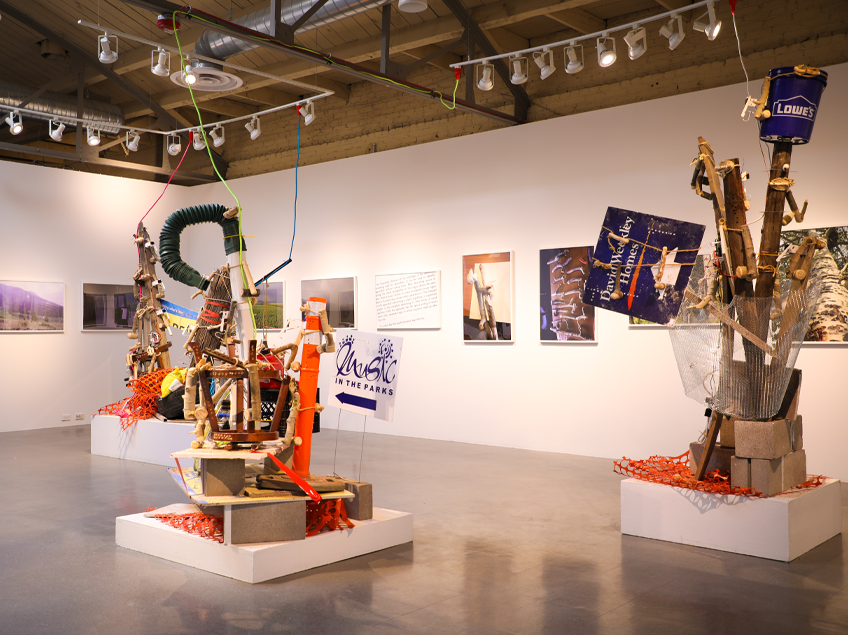 Art gallery with three large mixed-media sculptures using construction materials and signage. Framed abstract photos are displayed on a white wall.