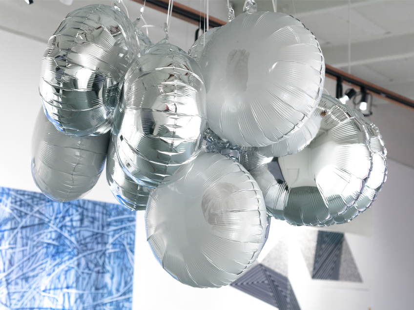 Gallery shot featuring a Mylar balloon installation suspended from the celing, with an abstract artwork in various blue hues displayed in the background.