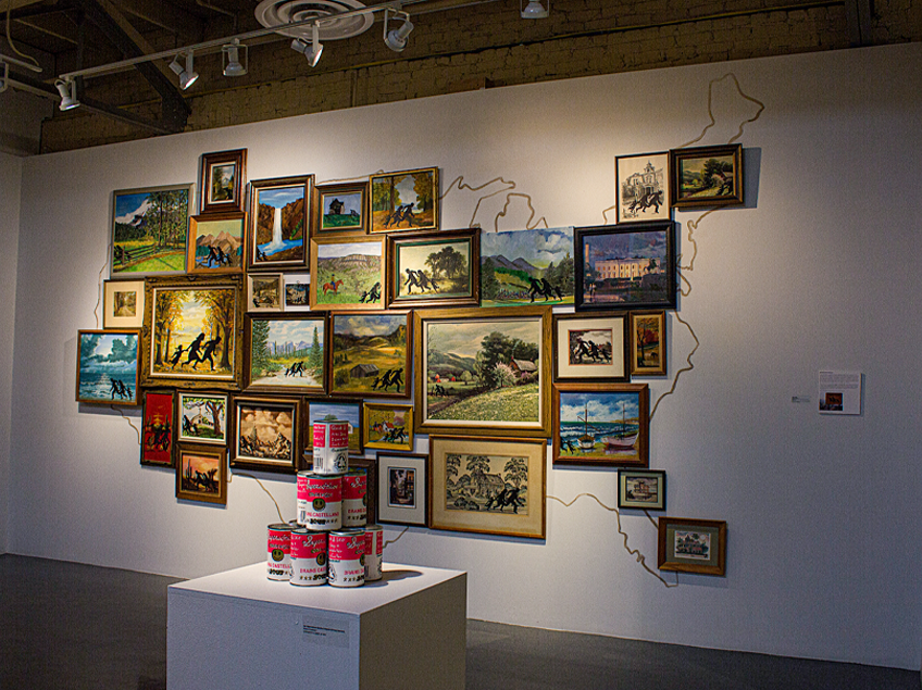 Art installation featuring framed paintings arranged in the shape of the USA and a pedestal with soup cans in the foreground.