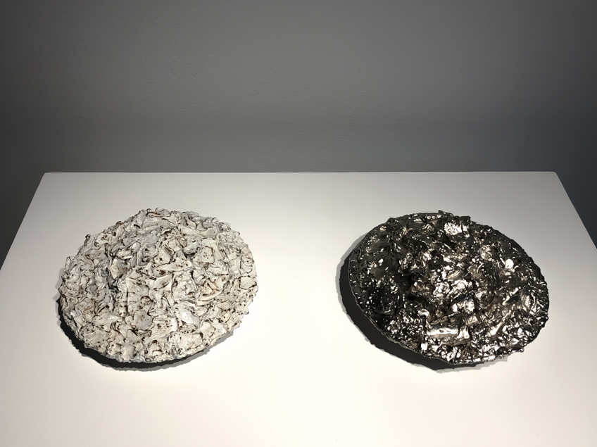 Two round, textured sculptures on a white surface; one light grey and rough, the other dark metallic and glossy.
