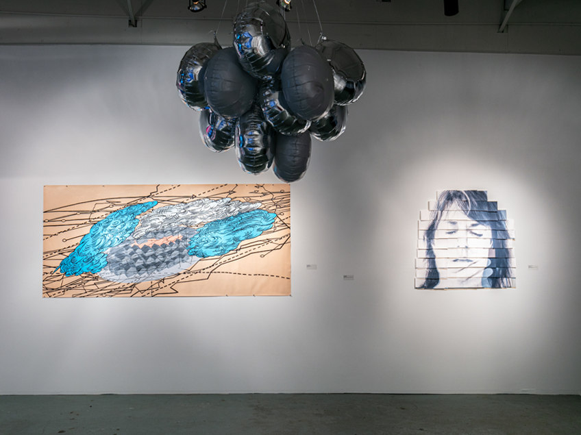 Gallery view featuring a Mylar balloon installation hanging from the ceiling. On the wall are two artworks: one an abstract painting in blues and tans with line work, and the other a face painting.