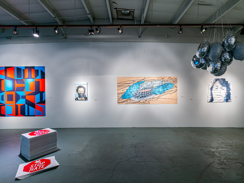 Gallery view, starting from the left: a bright blue, red, and purple painting next to a face portrait wearing a mask, followed by an abstract painting in blues and tans with line work, and another face painting. On the gallery floor is a pop art installation displaying the words 'Stop Hate'.