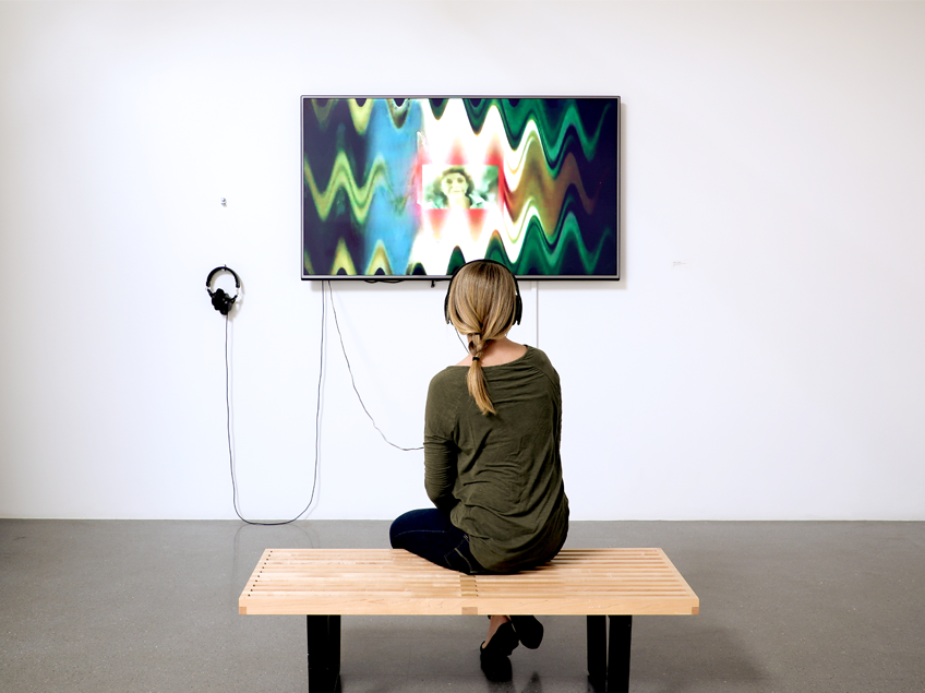 A person wearing headphones sits on a bench, watching an abstract video on a wall-mounted screen.