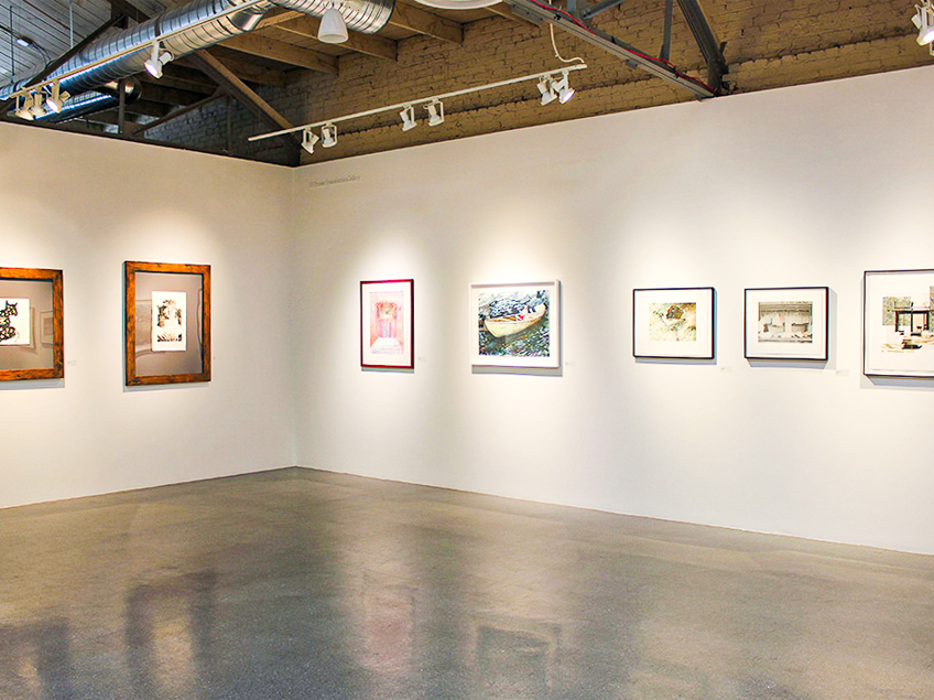 An art gallery corner with seven framed artworks on white walls, under spotlighting with exposed ceiling ductwork.