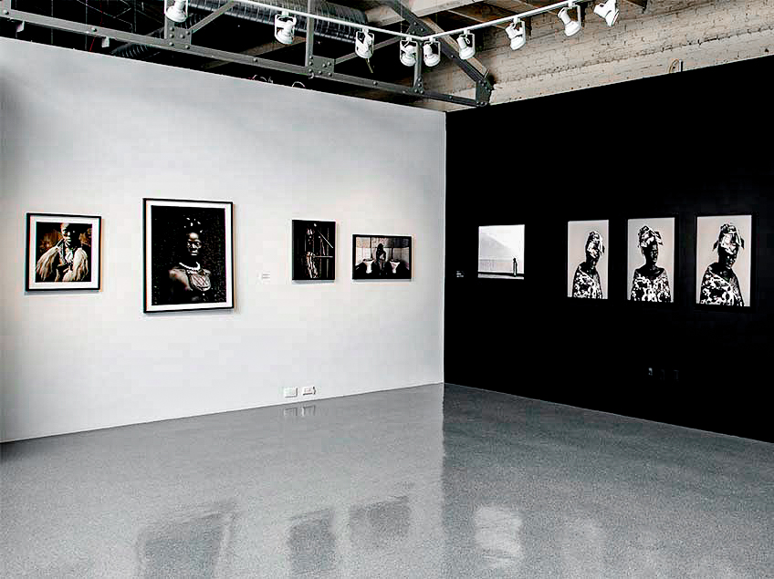 A gallery space with black-and-white photographs on white and black walls, featuring portraits and abstract art.