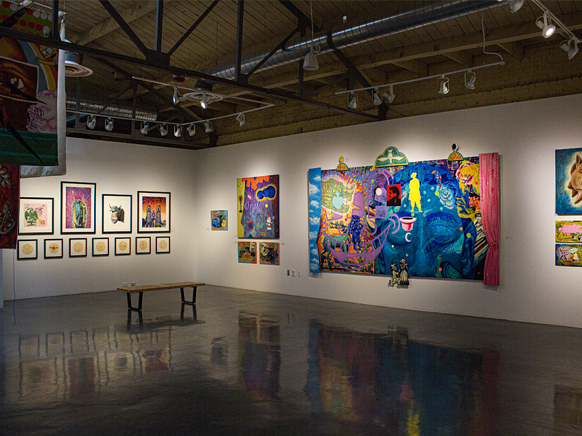 A gallery with colorful contemporary artworks on white walls and a wooden bench on a reflective floor.
