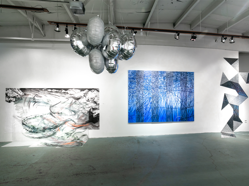 Gallery view featuring mylar balloon artwork suspended from the ceiling, with three abstract, colorful paintings displayed on the walls.